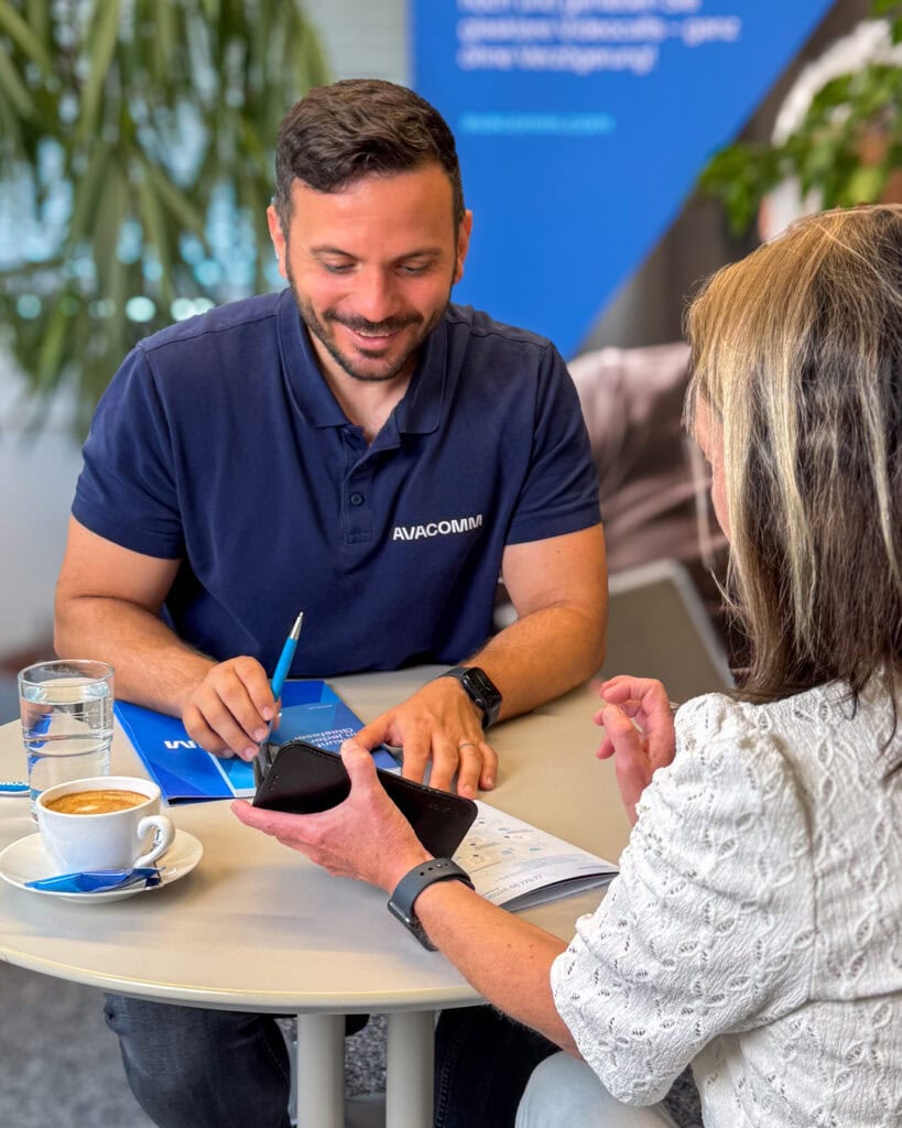 Ein Vertriebsmitarbeiter der Avacomm Systems GmbH (Glasfasernetzbetreiber) beantwortet die Fragen einer Kundin während der Glasfaser-Sprechstunde. Die Kundin zeigt auf ihr Smartphone und der Berater, ihr gegenüber sitzend, beantwortet lächelnd ihre Frage.