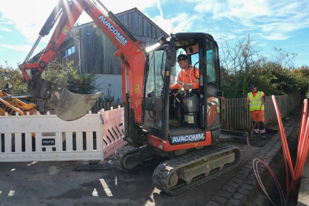 Bauarbeiten in Holzkirchen, ein kleiner Bagger trägt den Schriftzug Avacomm und zur rechten liegt die Speedpipe zum Verlegen bereit.