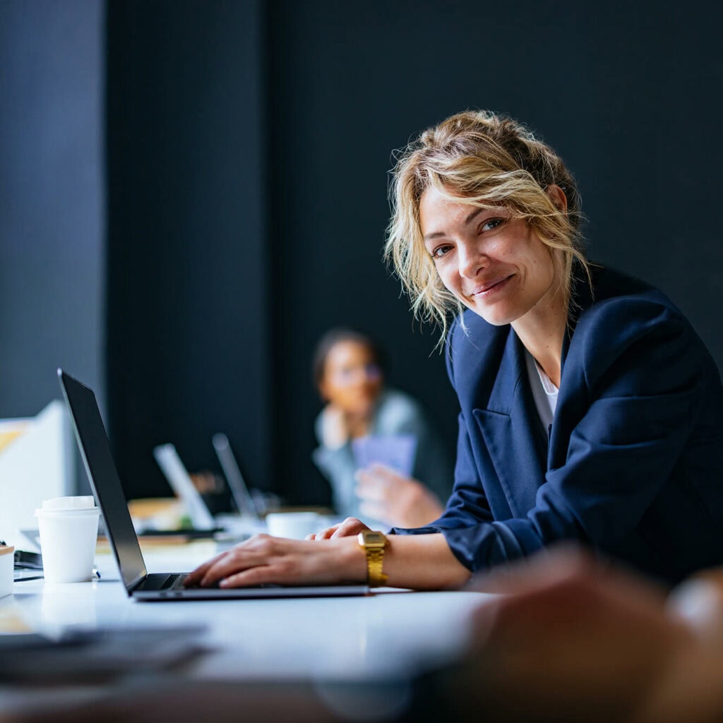 Frau am Laptop im modernen Büro am Schreibtisch mit einem Laptop sitzend, blickt in die Kamera – maximale Leistung und hohe Bandbreite durch zuverlässige Glasfaserverbindungen von AVACOMM