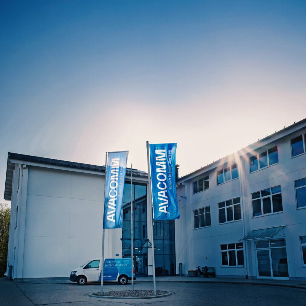 AVACOMM Firmenzentrale in Holzkirchen mit Lieferwagen im Avacomm-Branding und Unternehmensfahnen im Vordergrund – regionaler Anbieter für zukunftssichere Glasfaserlösungen.