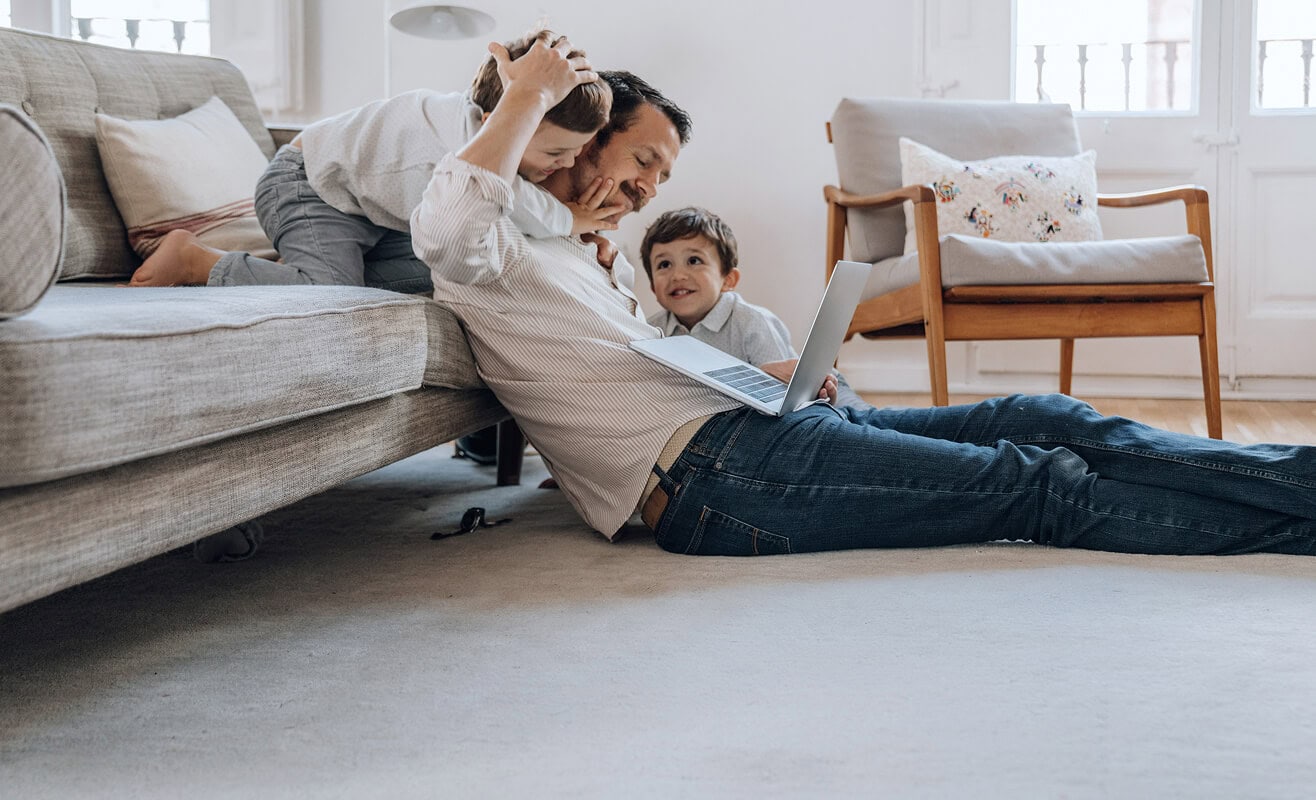 Vater spielt mit Kindern auf dem Boden, Laptop im Hintergrund – Glasfaser-Internet für modernes Familienleben in Mietwohnungen mit AVACOMM