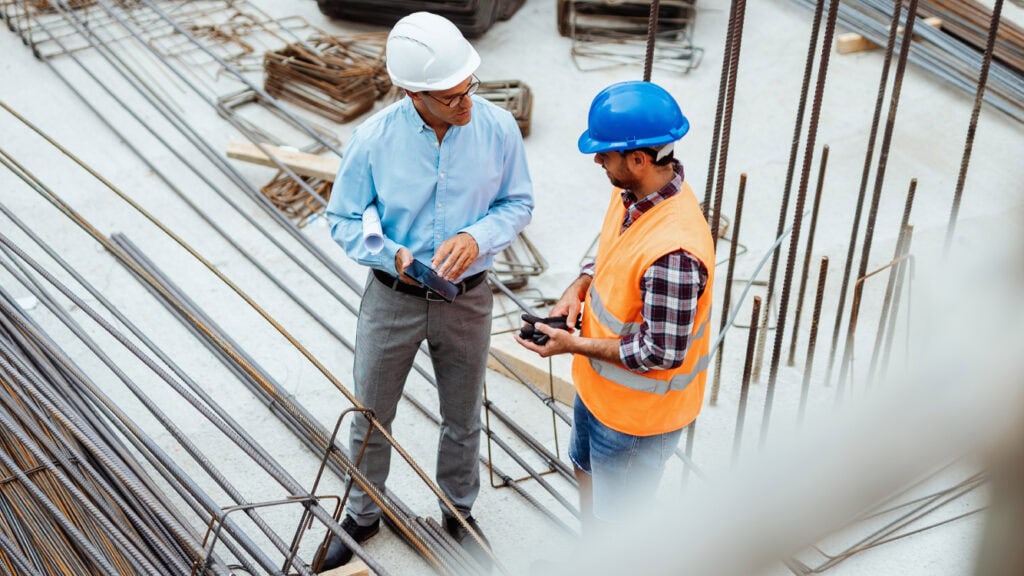 Architekt mit IPad in der Hand und Bauleiter mit Helm und Sicherheitsweste ausgestattet im Gespräch auf einer Baustelle im Rohbaustatus