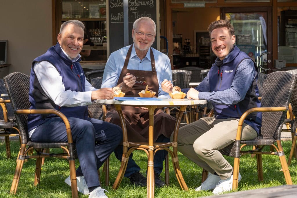 Herr Franz, Inhaber Eiscafe Franzetti sitzt im Sonnenschein auf der Terrasse, rechts und links von ihm Vertriebsmitarbeiter der AVAVACOMM. Gemeinsame Eisverkostung.